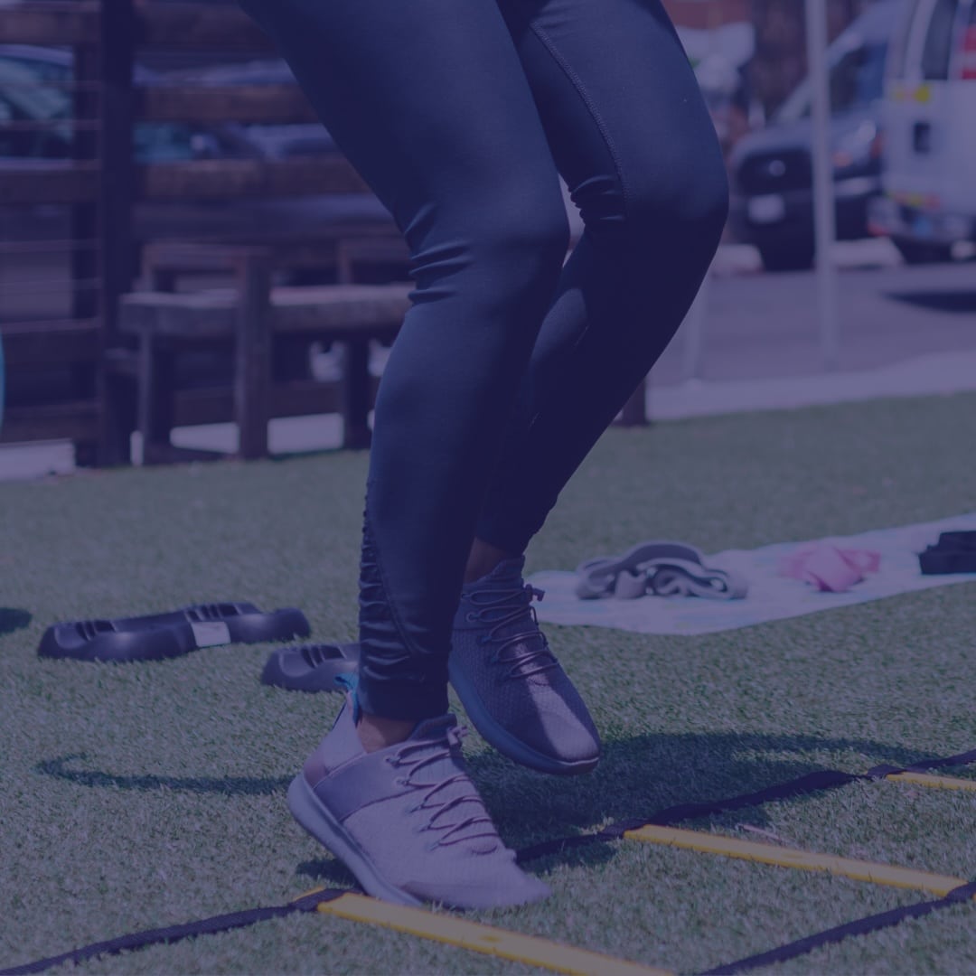 White House Sports Psychology – Featured – semi-private-training-featured Person wearing dark leggings and gray sneakers is doing ladder drills on artificial turf outdoors, with exercise equipment and vehicles visible in the background.