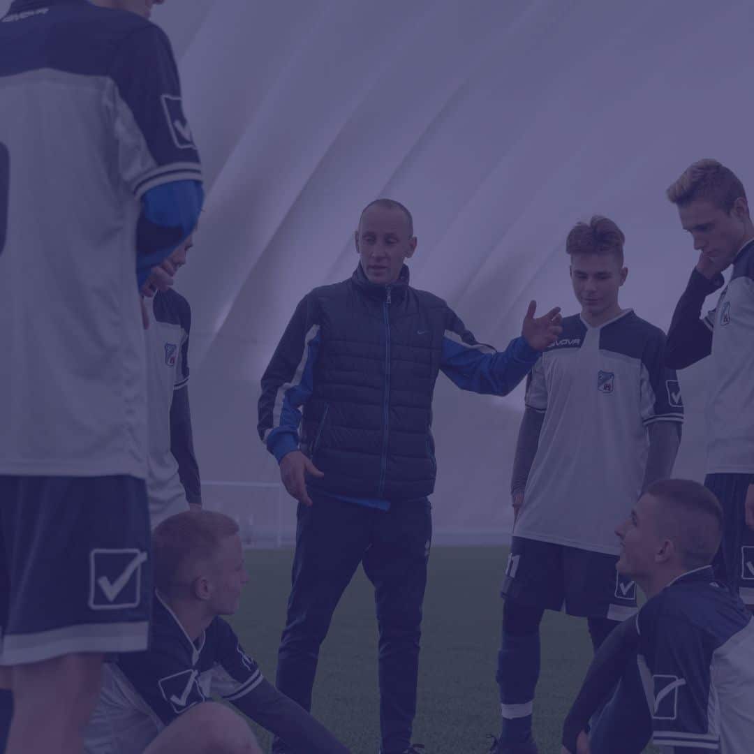 mental-toughness-more-than-a-buzzword-featured A coach stands and gestures while giving instructions to a group of young soccer players gathered around him on an indoor field.